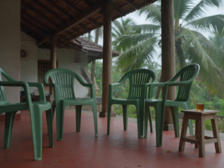 Four empty plastic chairs on a Kerala house veranda with red oxide floor and a tea glass on a low stool